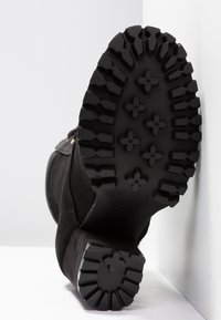 Semelle noire de bottine à talon avec une bande de roulement profonde comportant des motifs en caoutchouc en forme de fleurs et géométriques sur fond blanc.