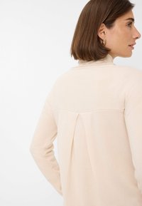 Un pull beige clair à manches longues, avec un détail de plis à l'arrière et une texture douce. Il présente un col standard.