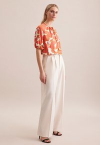 Korte mouwen oranje en witte bloezen met een ronde halslijn, gecombineerd met hoge witte broeken en zwarte haksandalen.