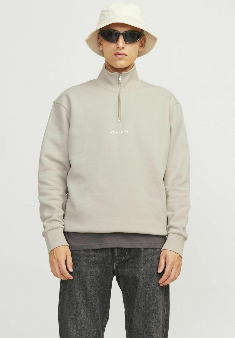 Light gray zip-up sweatshirt with a high collar, featuring a small logo. Worn over a dark layer, paired with dark jeans and a bucket hat.