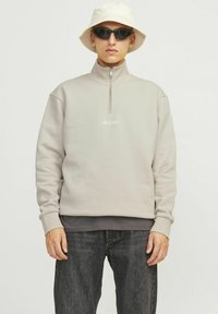 Vyras dėvi smėlio spalvos megztinį su ketvirčio užtrauktuku, juodus saulės akinius, baltą kibiro formos skrybėlę ir tamsiai pilkus džinsus, stovintis priešais paprastą foną.