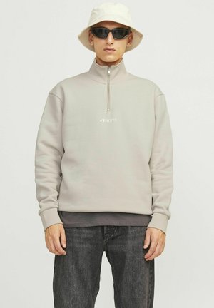 Helles graues Zip-Up-Sweatshirt mit hohem Kragen, ausgestattet mit einem kleinen Logo. Trägt man über einer dunklen Schicht, kombiniert mit dunklen Jeans und einem Bucket Hat.