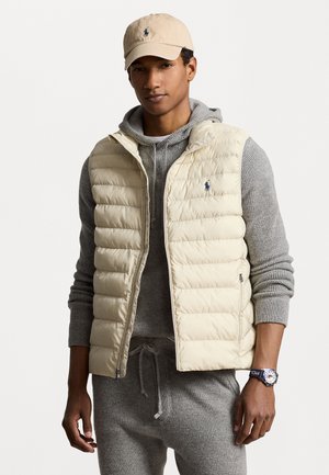 Puffervest i lys beige, med horisontal quilt og en liten blå logo. Grå strikket hettegenser under og grå joggebukser synlige.