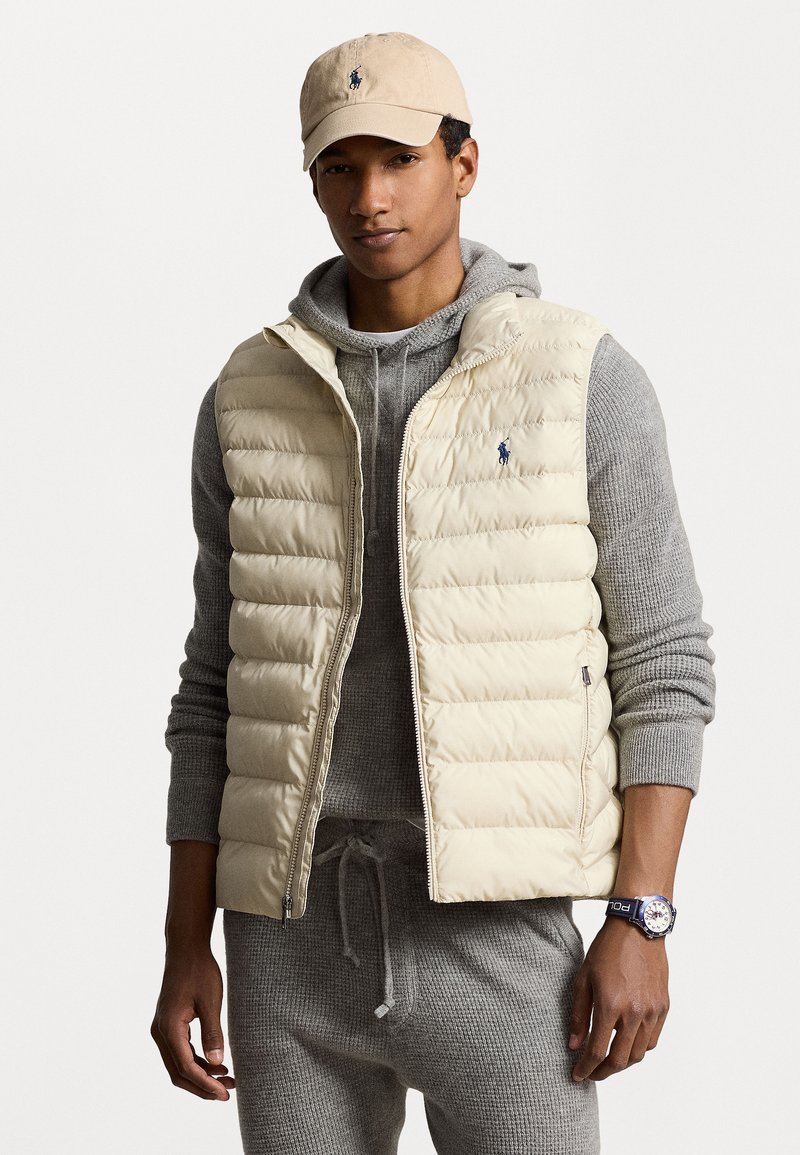 Gilet en duvet beige clair, avec un matelassage horizontal et un petit logo bleu. Sweat à capuche en tricot gris en dessous et pantalon de survêtement gris visible.