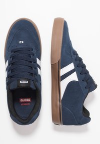 Navy blue suede sneakers with white side stripes, black interior, and brown rubber sole. Stylish design features laces and subtle branding.
