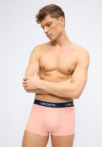 Άνδρας χωρίς πουκάμισο με κοντά σγουρά μαλλιά ποζάρει σταυρώνοντας τα χέρια, φορώντας ροζ ανοιχτό Lacoste μποξεράκια με navy μπλε ζώνη στη μέση.