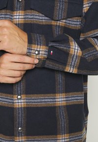 Camicia di flanella a quadri blu navy e senape con chiusura a bottoni a pressione e due tasche sul petto. Presenta un tessuto testurizzato e una piccola accentuazione del logo.