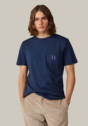 Young person with shoulder-length brown hair wearing a navy blue T-shirt with chest pocket and beige corduroy pants, hands in pockets.