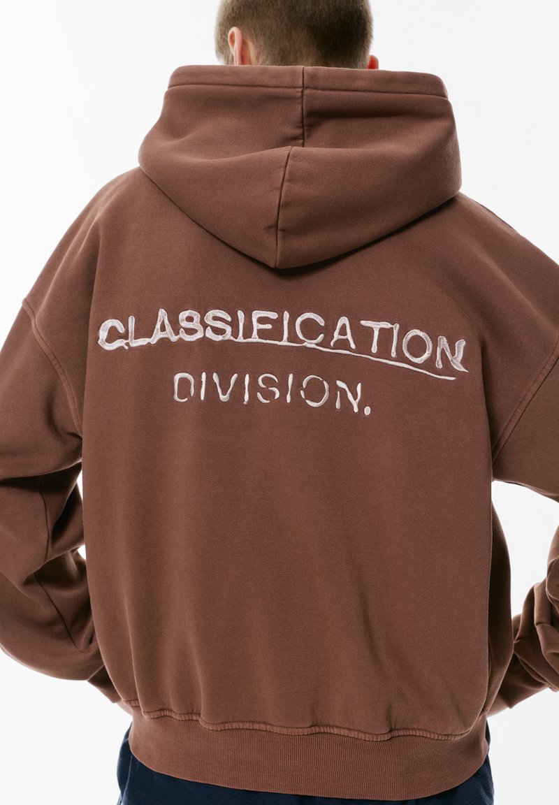 Hnedá mikina s kapucňou s veľkým vyšitým nápisom "CLASSIFICATION DIVISION" na chrbte. Mäkká tkanina, voľný strih a nastaviteľná kapucňa.
