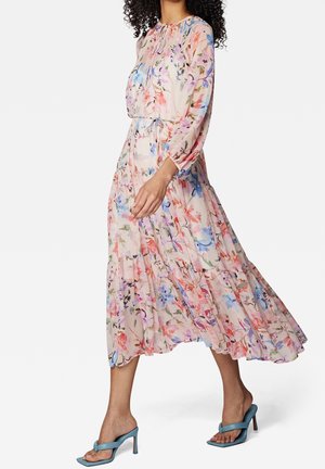 Sukienka midi w kwiaty z przezroczystej tkaniny, z długimi rękawami, falbanami i jasnoróżowym tłem. Noszona z niebieskimi sandałami na obcasie.