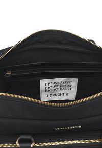 Borsa nera con cerniera dorata, dotata di interno foderato e di un'etichetta con la scritta ripetuta "I MUST RESIST".