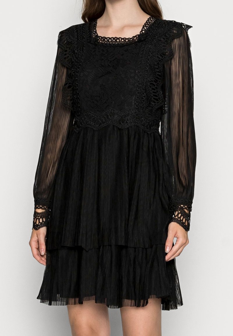 Femme portant une robe noire à manches longues avec des détails en dentelle sur le corsage, des manches transparentes plissées et une jupe plissée à plusieurs niveaux.