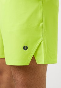 Pantalones cortos verde lima con abertura lateral y pequeño logo negro encima de la rodilla en la pierna desnuda de una persona.