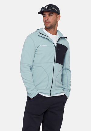 Man draagt een lichtblauwe fleecevest met ritssluiting, zwarte borstzak en zwarte broek, handen in de zakken, zwarte pet met zonnebril erop.