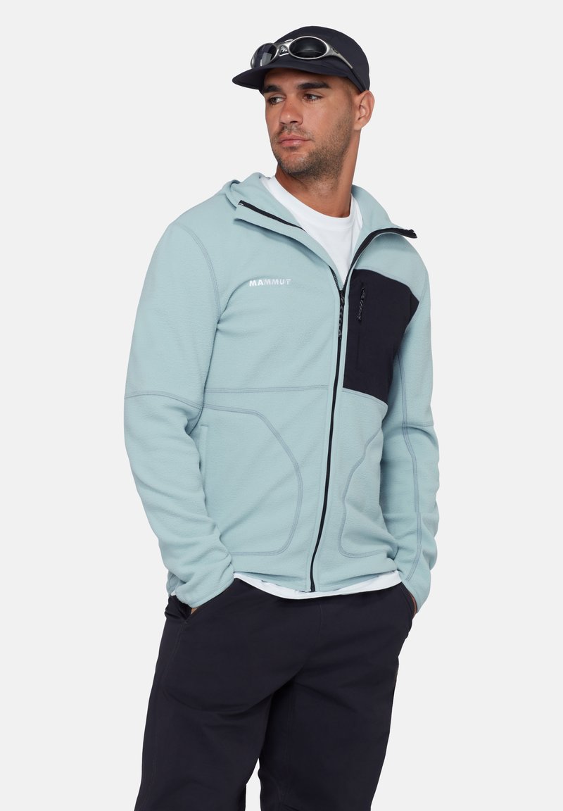 Homme portant une veste polaire à fermeture éclair bleu clair avec une poche poitrine noire et un pantalon noir, mains dans les poches, casquette noire avec des lunettes de soleil posées dessus.