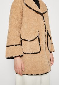 Brązowy, teksturowany futrzany płaszcz z czarną lamówką, dużymi przednimi kieszeniami i kołnierzem typu szal; rękawy o długości do połowy ramienia z kontrastującymi detalami przeszyć.