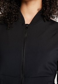 Kvinde iført en sort jakke med lynlås og ribbet krave, med krøllet hår synligt omkring skuldrene.