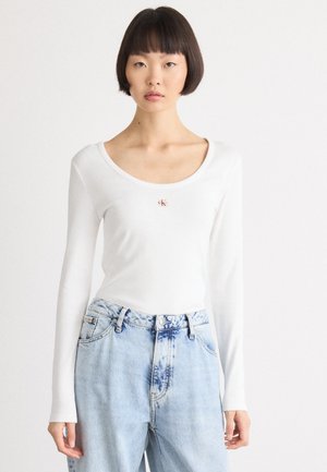 Hvit langermet topp med rund hals, med en liten rød logo. Kombinert med lyseblå høytlivs denimjeans med tie-dye detaljer.