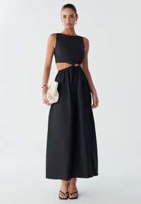 Model in een mouwloos, zwart maxi-jurk met een centrale uitsnijding en geknoopte details, terwijl ze een kleine crèmekleurige tas vasthoudt en minimale accessoires draagt.