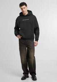 Svart huvtröja med texten "STARTED FROM THE BOTTOM" i vitt, framfickor, matchad med urtvättade bruna jeans och svarta skor.