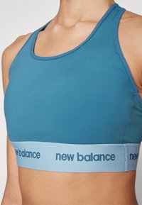Modra športna nedrček z gladkim, prilegajočim se dizajnom in širokim svetlo modrim pasom, na katerem je v temno modri barvi napis "new balance".