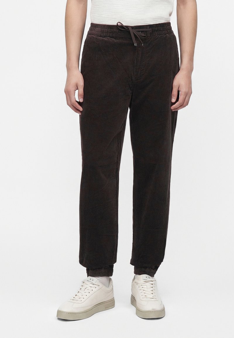 Homme portant un pantalon en velours côtelé marron foncé avec taille à cordon et des baskets blanches sur un fond uni.