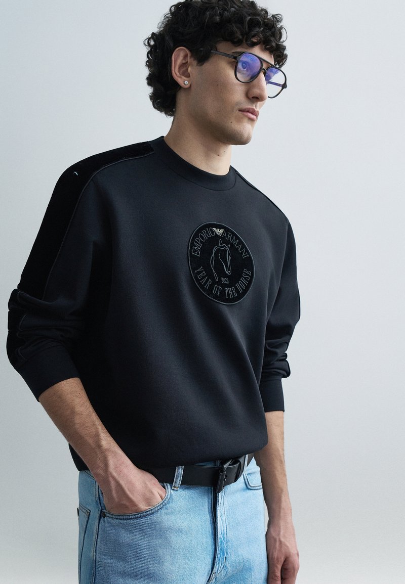 Melns sporta krekls ar apaļu emblēmu, kurā attēlots zirga dizains un teksts. Valkāts kopā ar gaiši ziliem džinsiem, kas demonstrē brīvu piegriezumu un tekstūru.