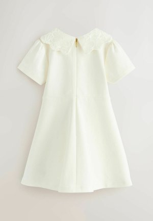 Balta kleita ar īsām pūkām, viļņotu izšūtu apkakli un aizmugurē esošu rāvējslēdzēju. A-veida forma un gluda, mīksta tekstūra.