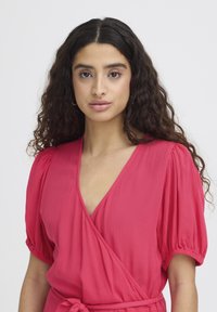 Jeune femme aux longs cheveux bouclés et foncés, portant une robe portefeuille rose à manches bouffantes et taille nouée, face à l'avant sur un fond blanc.