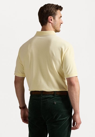Tricou polo galben cu mânecă scurtă dintr-un material texturat, prezentat din spate, asortat cu pantaloni din corduroy verde închis și o curea maro.