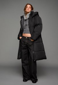 Černý prošívaný dlouhý kabát přes šedý pletený crop top a tmavé široké džíny. Obsahuje kapuci, stahovací šňůrky a texturovaný úplet. Černé boty dol completing celý vzhled.