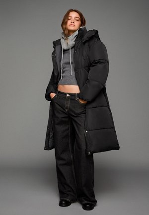 Zwarte gewatteerde lange jas over een grijze gebreide crop top en donkere wijde jeans. De jas heeft een capuchon, trekkoord en een gestructureerde breiwerk. Zwarte laarzen maken de look af.