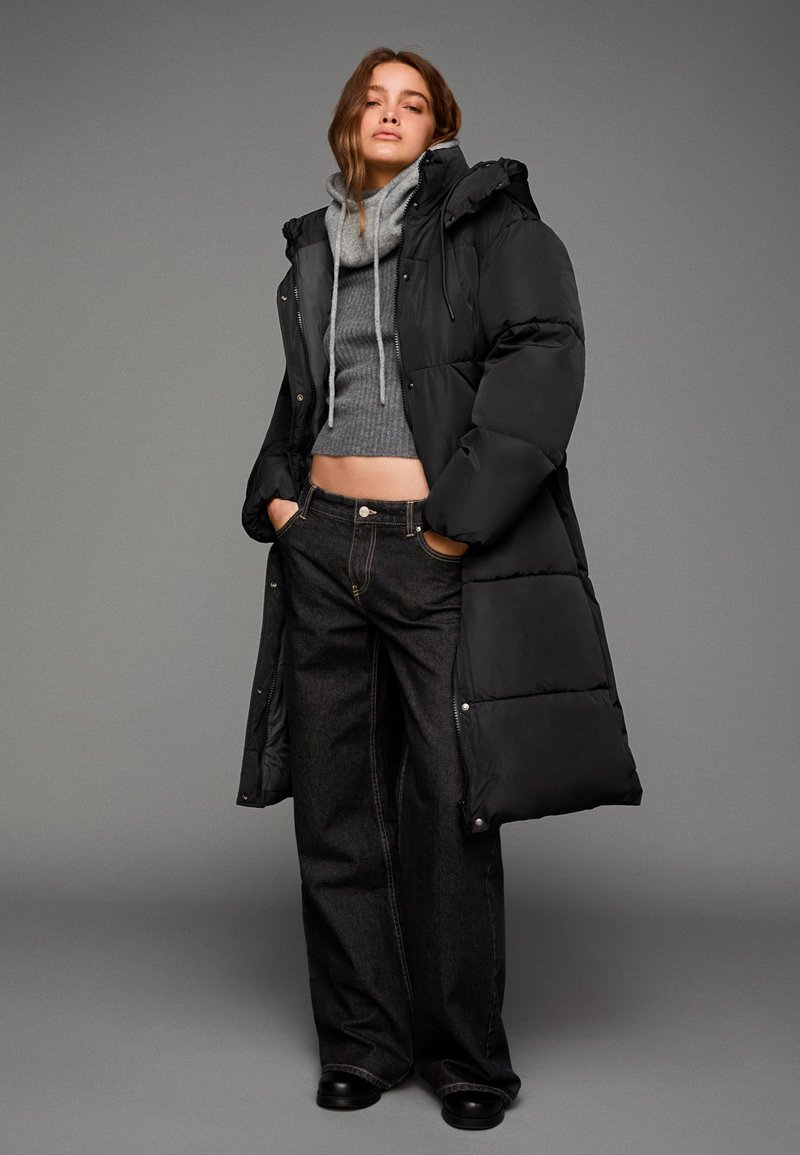 Černý prošívaný dlouhý kabát přes šedý pletený crop top a tmavé široké džíny. Obsahuje kapuci, stahovací šňůrky a texturovaný úplet. Černé boty dol completing celý vzhled.