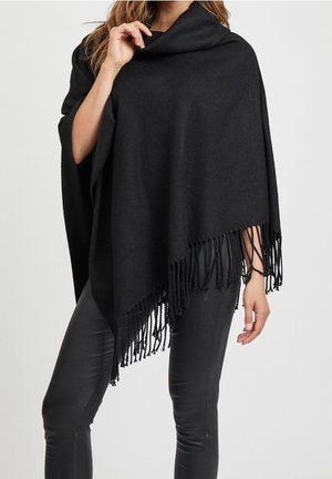 Kobieta ubrana w czarny poncho z frędzlami na czarnych skórzanych spodniach, trzymająca kołnierz jedną ręką na szarym tle.