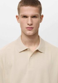 Béžové polo tričko s klasickým límcem, které má dvoubotové zapínání a hladký povrch; jednoduchý, minimalistický design.