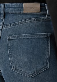 Close-up de um bolso traseiro de umas calças de ganga azuis com bordas costuradas e uma etiqueta de couro castanho com a inscrição "Anno 1995 / Blue Denim ONLY."