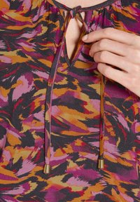 Bunt gemusterte Bluse mit langen Ärmeln, V-Ausschnitt, Kordelzügen mit goldenen Beschlägen und einem auffälligen abstrakten Muster in Lila, Orange und Schwarz.
