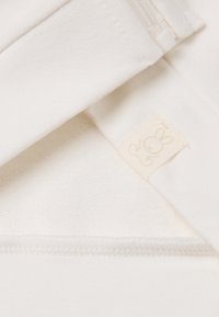 Tessuto bianco morbido con un'etichetta piccola che mostra un semplice contorno di un coniglio, evidenziando le cuciture ripiegate e un dettaglio della cerniera bianca.