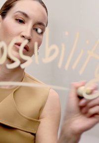 Top beige senza maniche con un design strutturato. Una mano scrive 'possibilità' su una superficie trasparente con un pennarello.