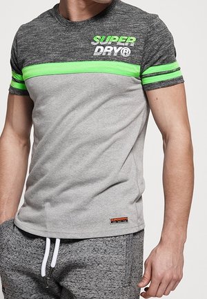 Mężczyzna w szarej i ciemnoszarej koszulce z neonowozielonymi paskami i logo "SUPER DRY", w połączeniu ze szarymi spodniami dresowymi z białymi troczkami.