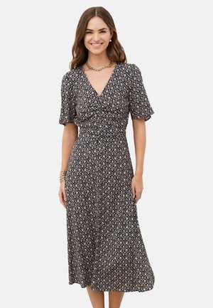 Femme souriante portant une robe midi noire et blanche à motifs, manches courtes et col en V, accessoirisée avec des bijoux dorés.
