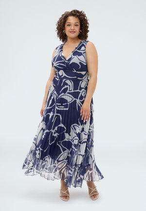 Vrouw met krullend haar, gekleed in een mouwloze navyblauwe en witte bloemen geplooide maxi-jurk en witte sandalen met bandjes.