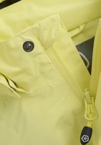 Ljusgul vattentät jacka med synlig dragkedja och tryckknapp, med texturerad tyg och en dragflik för enkel fastsättning.