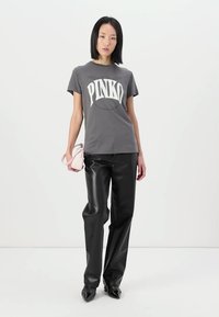 Camiseta de algodón gris con gran logo blanco "PINKO", combinada con pantalones de cuero sintético negros y sosteniendo un pequeño bolso rosa.