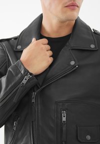 Giacca da moto in pelle nera con colletto ampio, cerniere sulle maniche e molteplici tasche con zip. Texture liscia con dettagli in metallo.