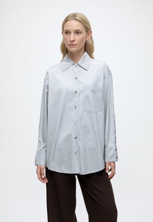 Ženska s svetlimi lasmi, oblečena v ohlapno svetlo sivo srajco z gumbi in temno rjave hlače, na enobarvni beli podlagi.