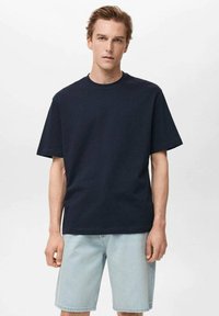 Tricou bleumarin cu mânecă scurtă din bumbac, cu guler rotund, purtat cu pantaloni scurți din denim deschis, având un croi lejer și un design minimalist.