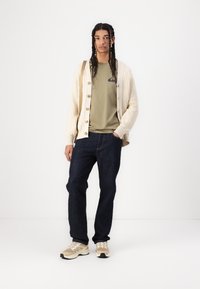 Beige knitted cardigan over an olive t-shirt, dark jeans, and beige athletic shoes with light accents. The outfit features buttons and a logo.