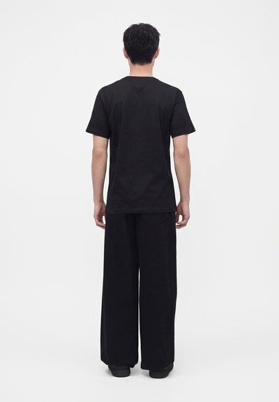 Homme debout, de dos, portant un t-shirt noir uni à manches courtes et un pantalon noir à jambes larges sur fond blanc.