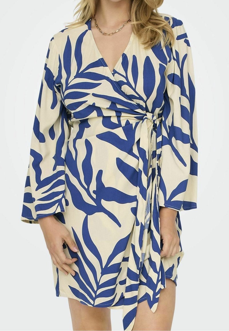 Femme portant une robe portefeuille beige avec de grands motifs de feuilles bleues et une ceinture latérale, associée à un collier chaîne en or.
